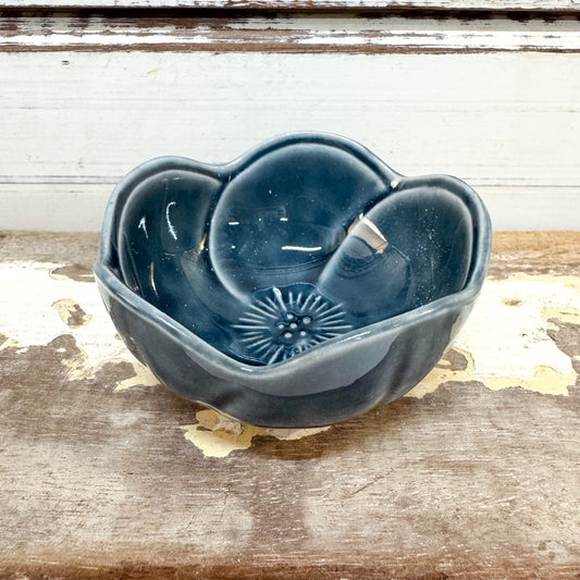 Concept Japan - Plum Petal Dish - Navy A small ceramic flower-shaped bowl in a dark blue-gray glaze, placed on a rustic wooden surface with a distressed white wall in the background.