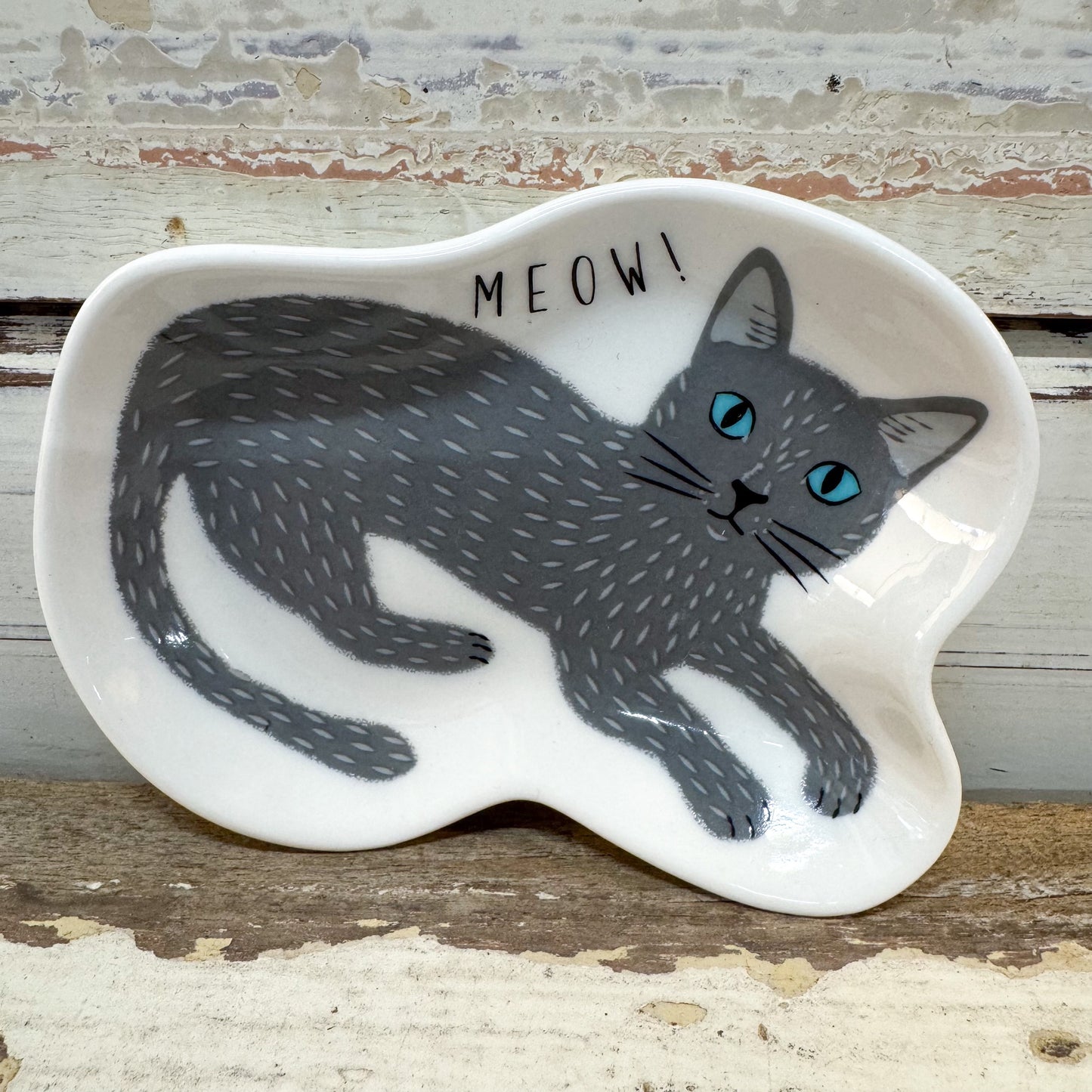 A white ceramic cat-shaped dish featuring a gray cat illustration with bright blue eyes and the word 'MEOW!' printed above it, set against a rustic distressed wood background. Concept Japan | Grey Cat Dish