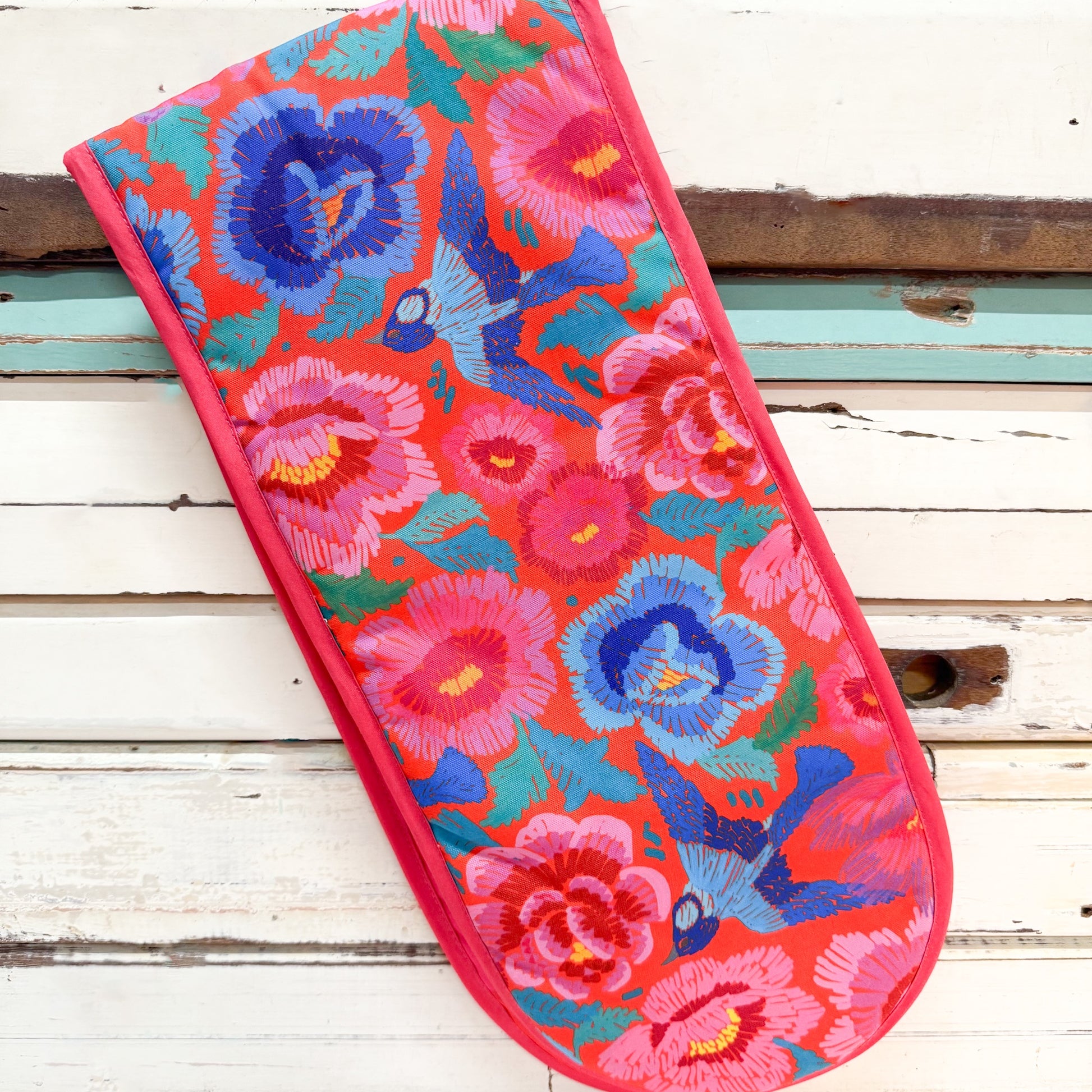 A rectangular quilted fabric item, likely a pillowcase or decorative cover, featuring a vibrant floral and bird pattern on a red-orange background. The design includes pink and blue flowers with green leaves and blue birds.