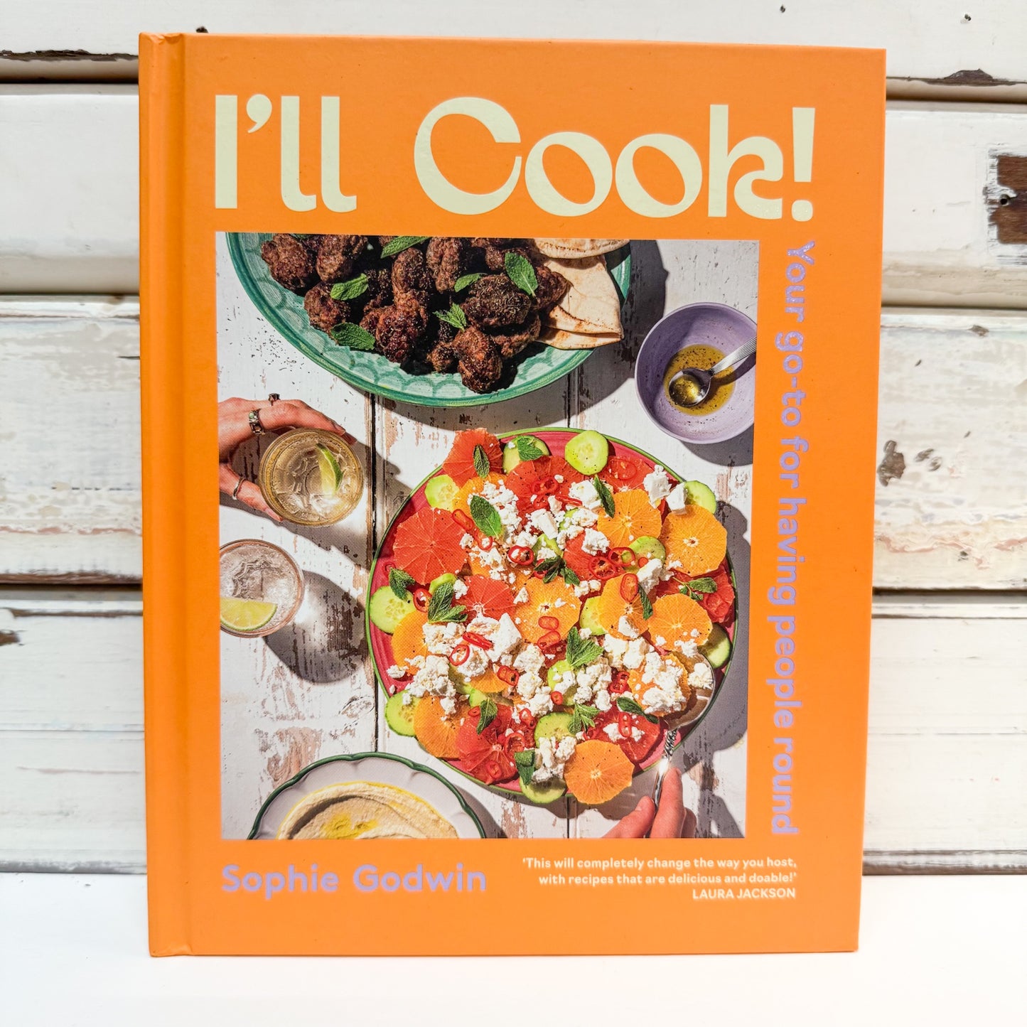 The book 'I'll Cook!' by Sophie Godwin, featuring a bright orange cover with a colorful photograph of a fresh salad and other dishes, set against a white wooden background.