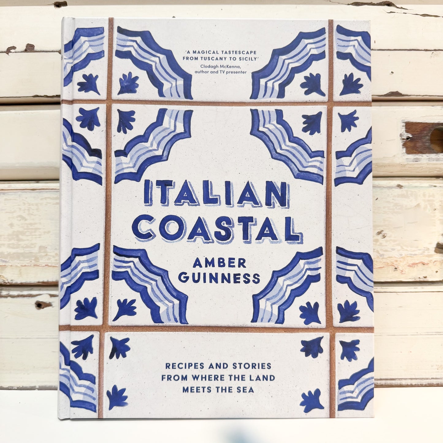 The book cover of 'Italian Coastal' by Amber Guinness, featuring a white background with blue Mediterranean-style tile patterns and brown grid lines, photographed flat on a rustic white wooden surface.
