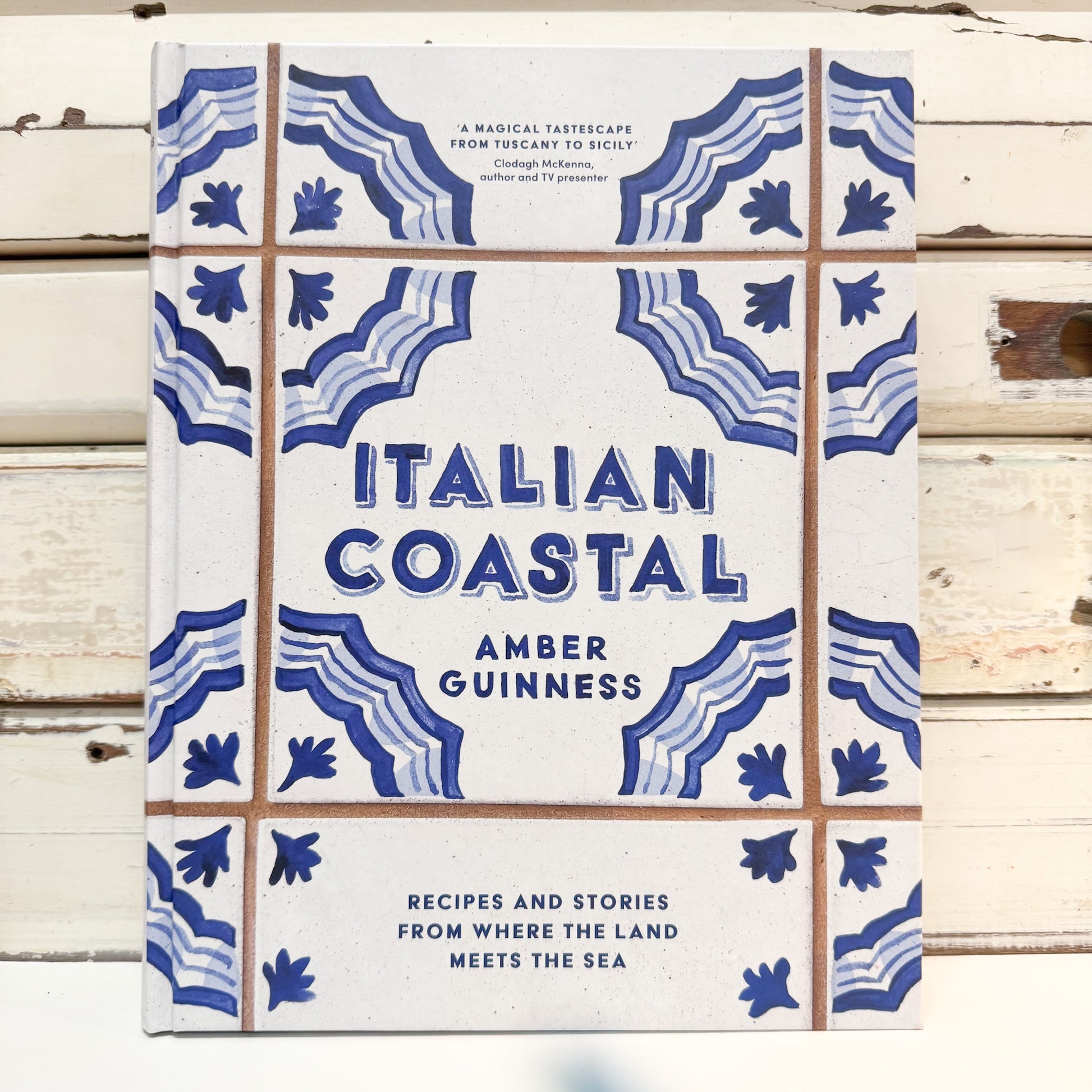 The book cover of 'Italian Coastal' by Amber Guinness, featuring a white background with blue Mediterranean-style tile patterns and brown grid lines, photographed flat on a rustic white wooden surface.