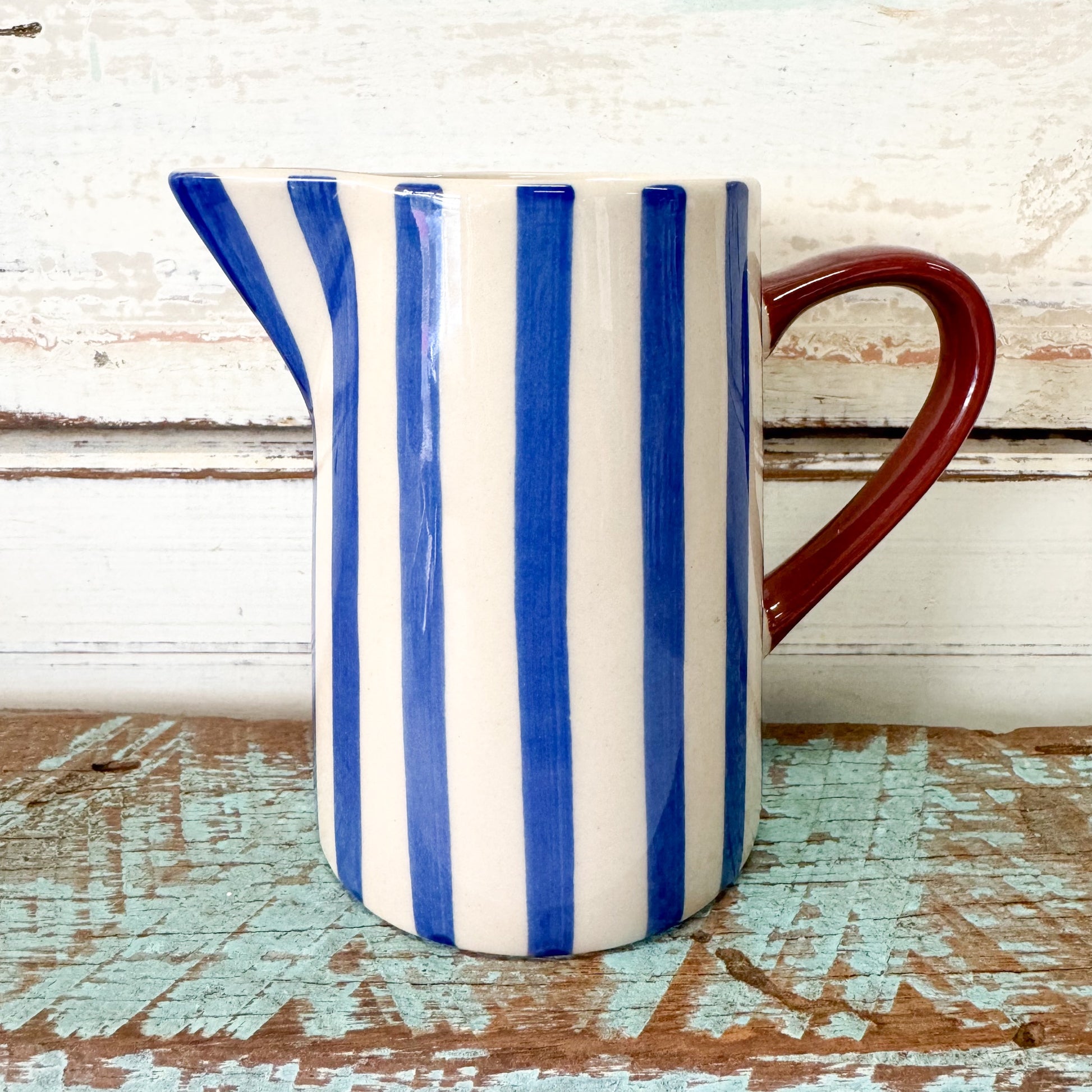 Ceramic pitcher with blue and white stripes and a red handle on a wooden surface. Jones & Co - Bloomie Blue Milk Jug