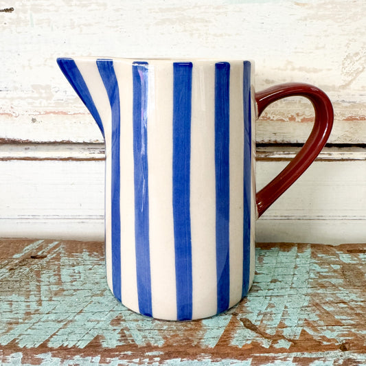 Ceramic pitcher with blue and white stripes and a red handle on a wooden surface. Jones & Co - Bloomie Blue Milk Jug
