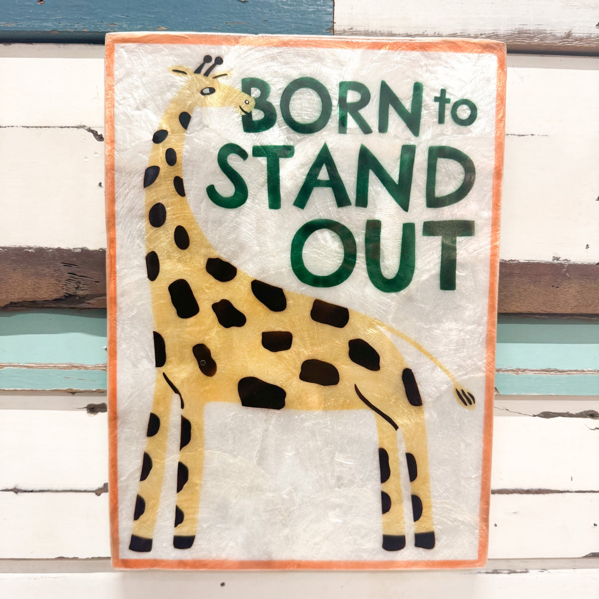 A rectangular decorative sign featuring a stylized giraffe and the phrase “BORN to STAND OUT” in dark green lettering, mounted against a distressed white wood plank background.