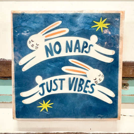 A square decorative wall sign with a distressed blue background featuring two white rabbits lying down with the text 'NO NAPS JUST VIBES' in blue, plus two yellow star accents, photographed against a white painted wood surface.