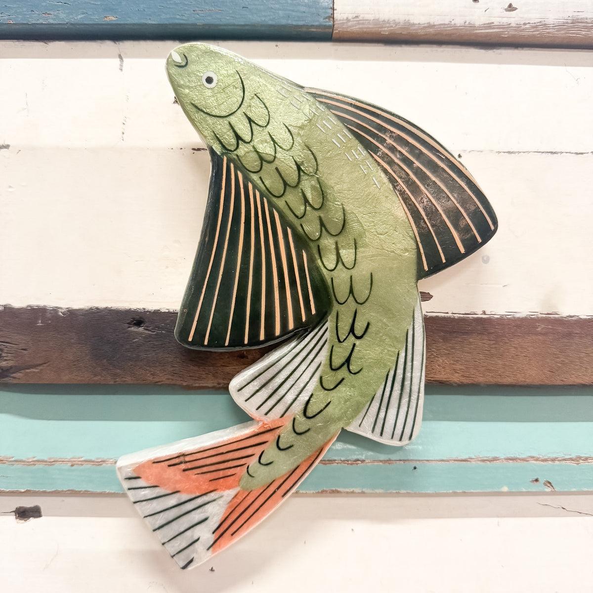 A hand-painted capiz shell fish wall hanging with a green body, black and gold pinstriped dorsal fin, white and black pinstriped vent fins, and orange and black striped tail, mounted on a rustic white and teal wooden plank background.