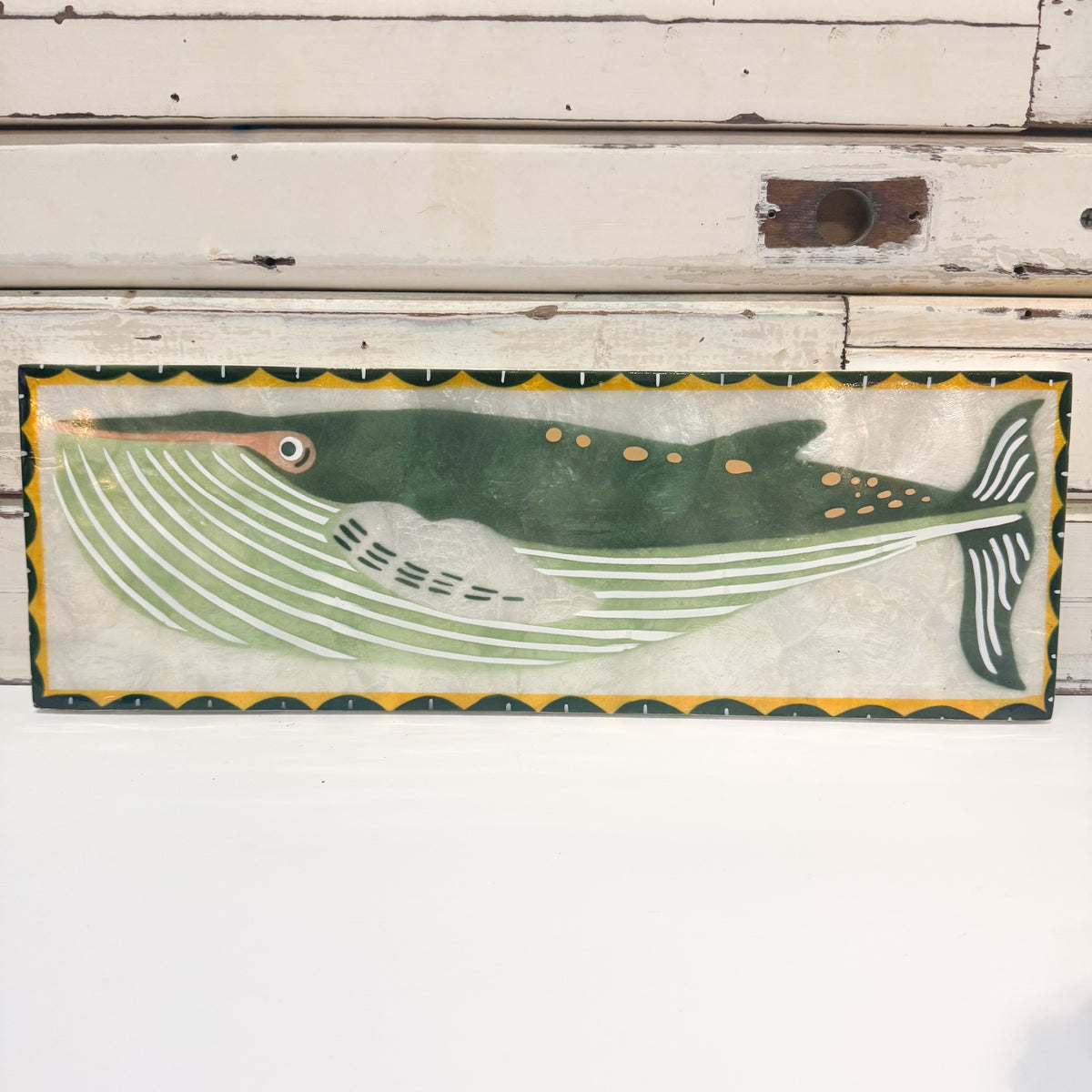 A rectangular hand-painted whale wall plaque featuring a green whale with white and light green striped details, set against a distressed white wood background.