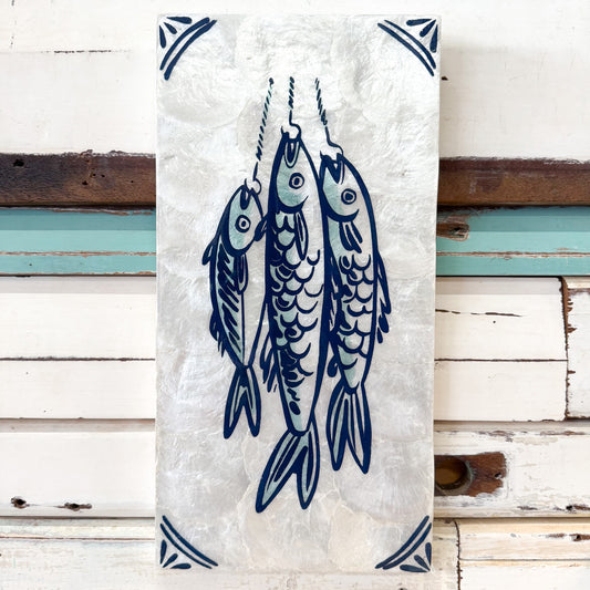 A rectangular white wall hanging or tile with a blue illustration of three fish hanging from hooks, bordered with blue corner motifs, displayed against a white wooden plank wall.
Jones & Co | Taverna Fish