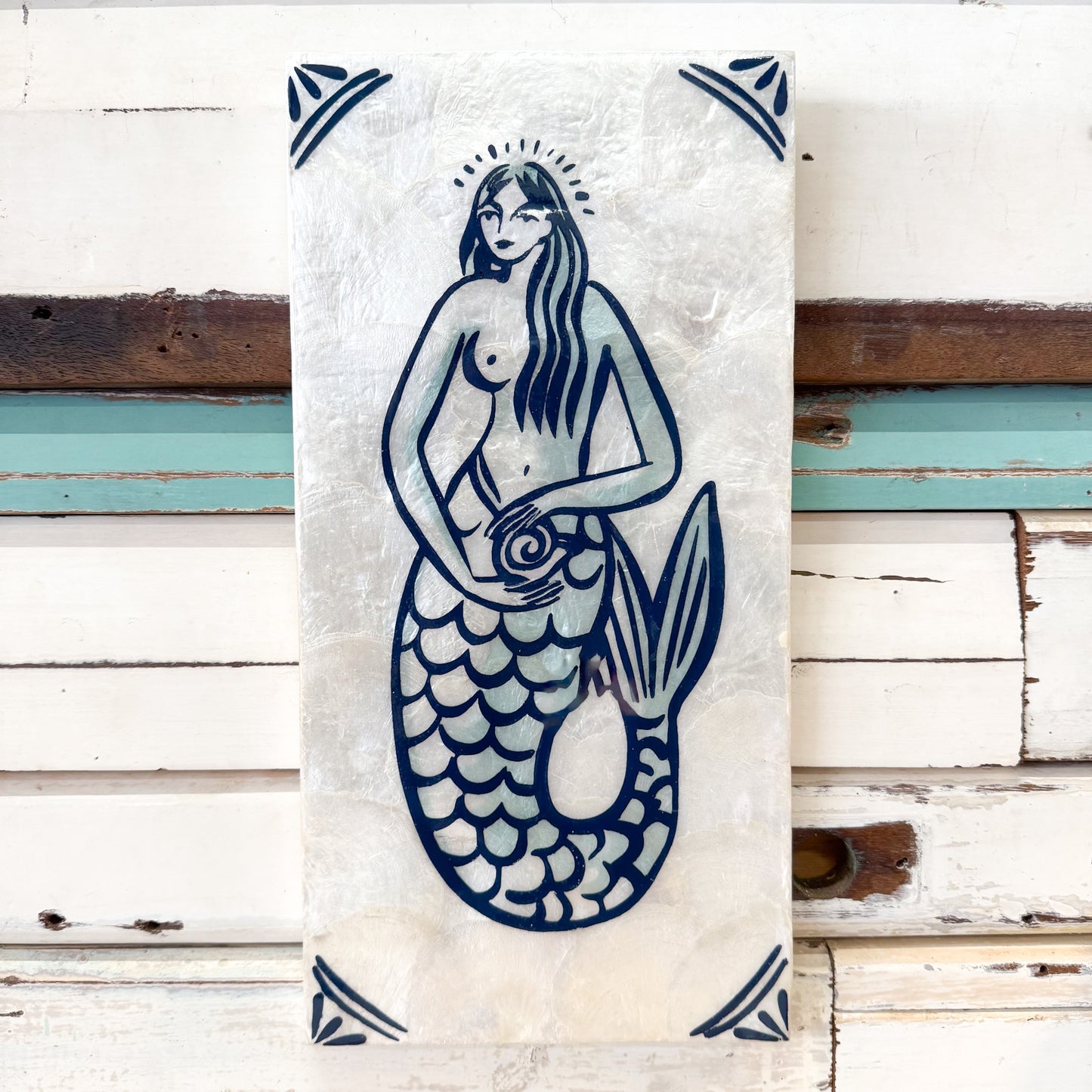 A vertical rectangular wall art piece featuring a blue mermaid illustration on a textured off-white background, displayed against distressed white wooden siding with weathered brown and teal paint. Jones & Co | Taverna Mermaid