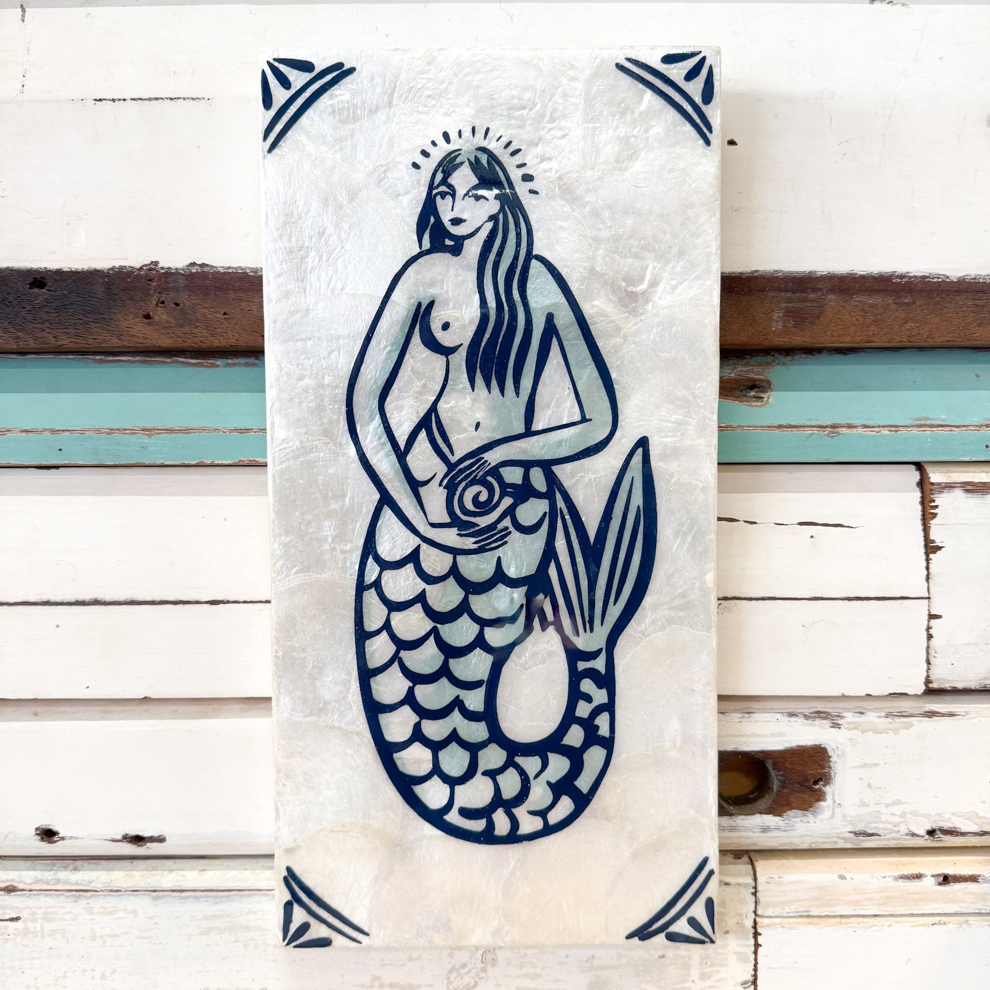 A vertical rectangular wall art piece featuring a blue mermaid illustration on a textured off-white background, displayed against distressed white wooden siding with weathered brown and teal paint. Jones & Co | Taverna Mermaid