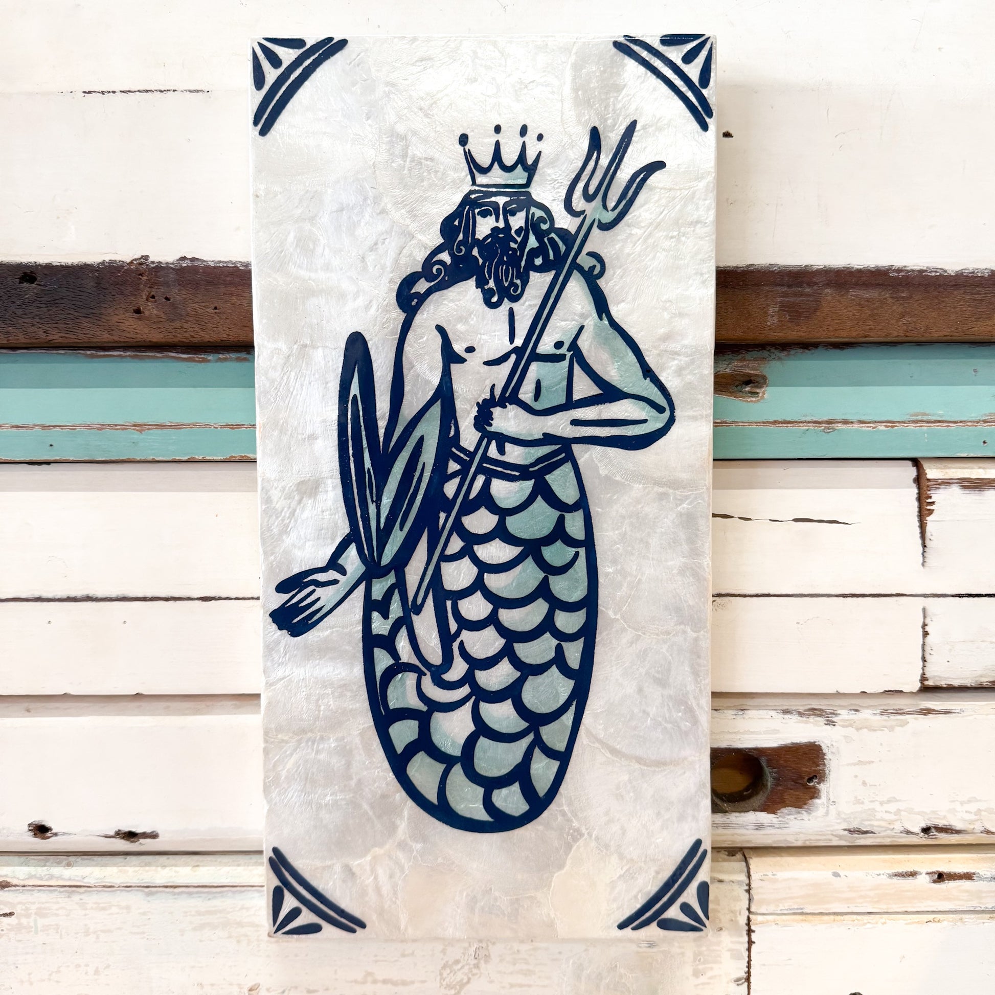 A vertical rectangular wall art print or tile featuring a stylized merman king with a crown, beard, trident, and shield in dark blue line art on a textured off-white background, displayed against a rustic white wood plank wall.
Jones & Co | Taverna Trident