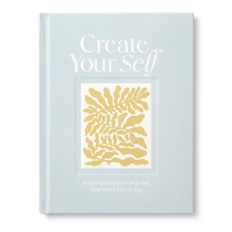 A blue hardcover journal with a title 'Create Your Self' and a small gold floral design on the cover, with the subtitle 'Simple Questions to Shape and Grow Every Part of You'.