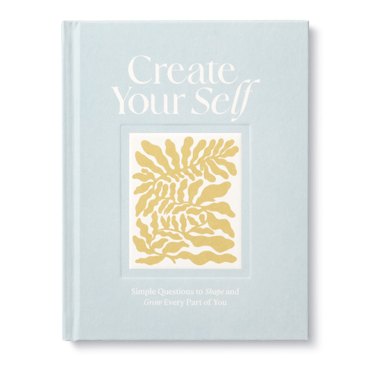 A blue hardcover journal with a title 'Create Your Self' and a small gold floral design on the cover, with the subtitle 'Simple Questions to Shape and Grow Every Part of You'.