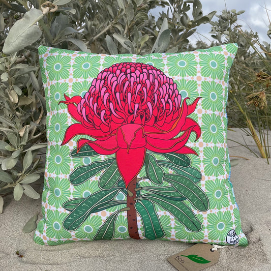 LOST AND FOUND ART CO Waratah 40cm Cushion Decorative pillow with a pink flower design on a green patterned background, placed outdoors.