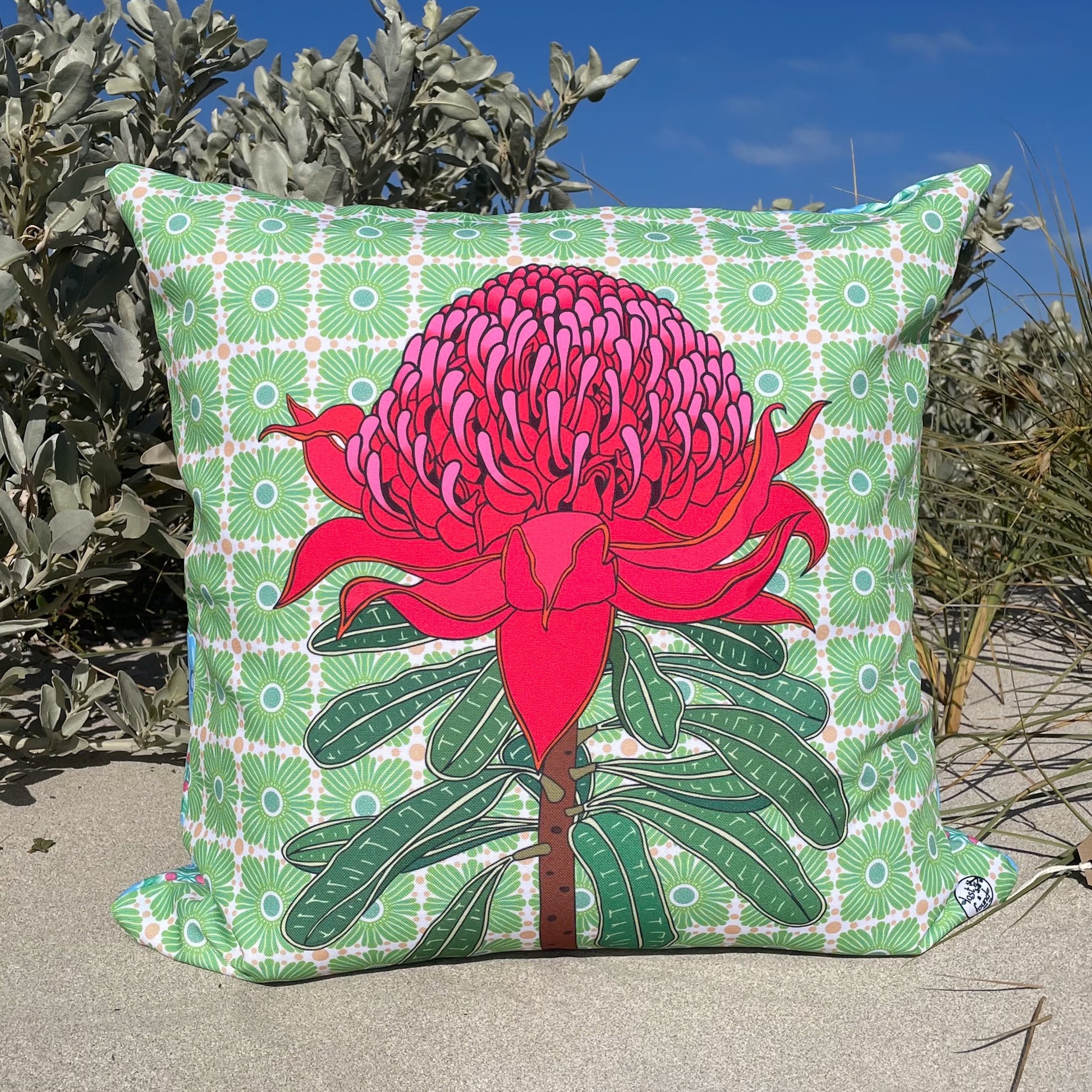Pillow with a red pink Waratah flower design on a green patterned background, LOST AND FOUND ART CO Waratah 60cm Cushion outdoors.