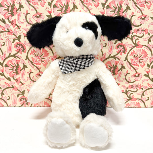 A plush puppy toy with black and white fur, wearing a black and white gingham bow tie, posed against a pink floral patterned background.
