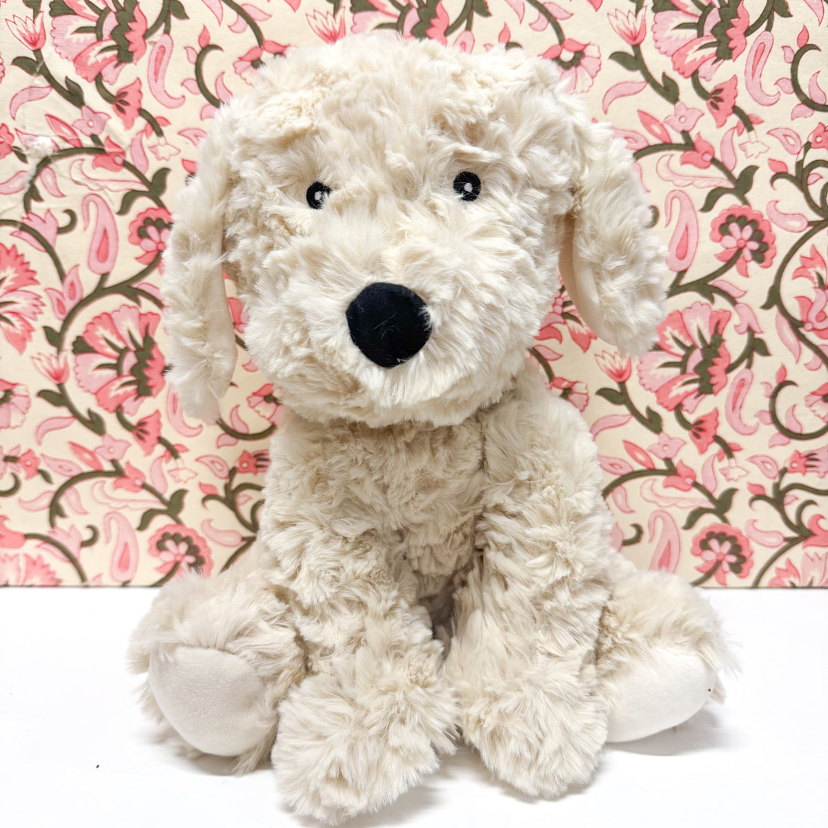A fluffy cream-colored plush dog toy with a black nose and black eyes, sitting in front of a pink and green floral patterned backdrop.