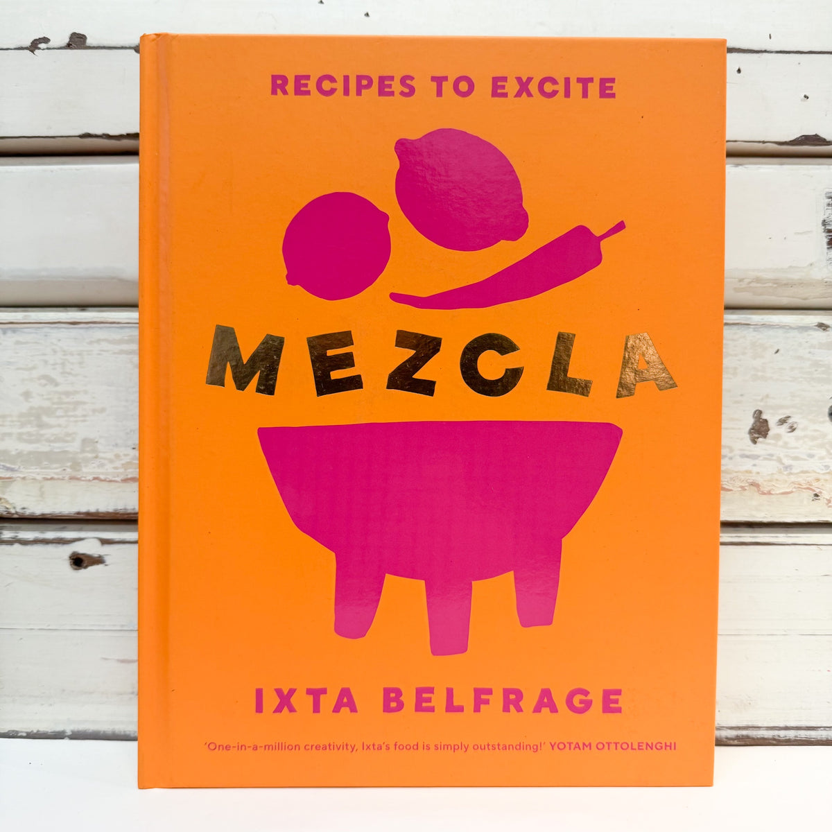 The front cover of the book 'Mezcla: Recipes to Excite' by Ixta Belfrage, featuring a bright orange background with bold magenta illustrations of lemons, a chili pepper, and a mortar and pestle, and the title in large metallic gold letters, set against a white wood plank background.