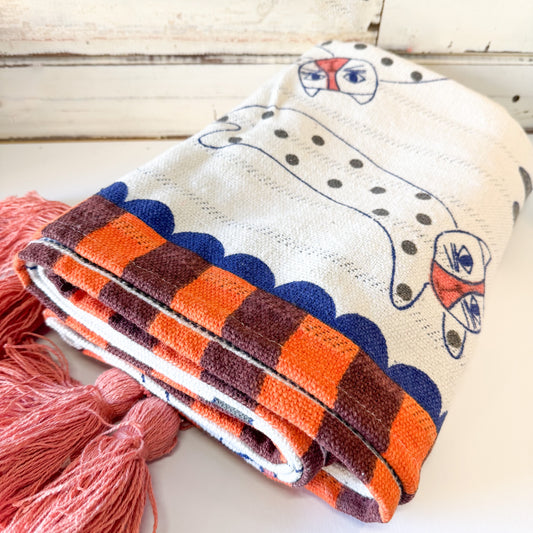A folded cotton blanket with a cream base, featuring a dachshund pattern, a blue scalloped border, and orange and brown checkered stripes, styled with pink tassels on a white wooden surface.