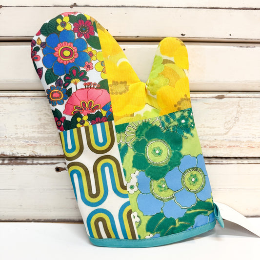 A single fabric oven mitt with a colorful retro patchwork floral and geometric print, displayed against a rustic white wooden plank background.