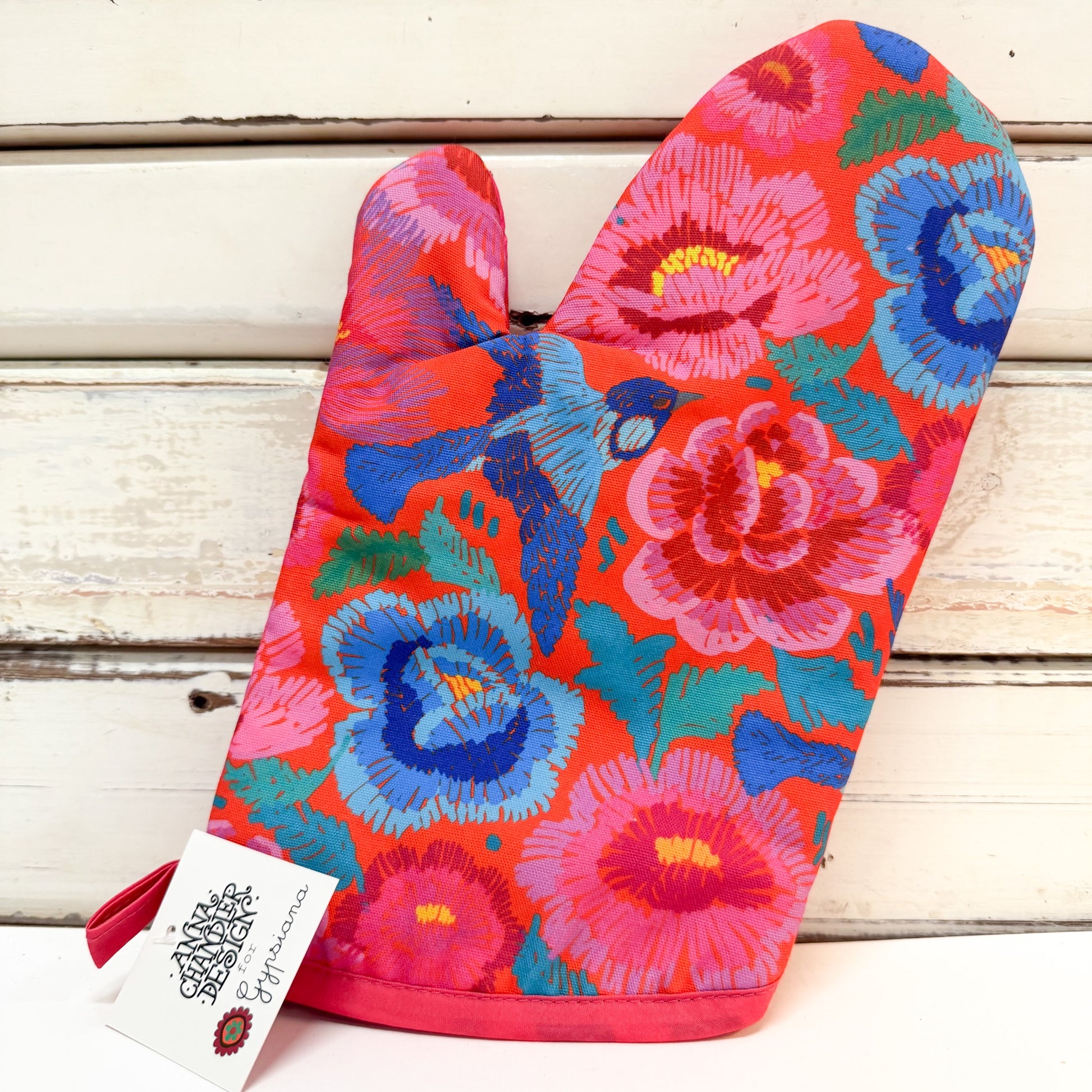 A single oven mitt with a vibrant floral and bird pattern in pink, blue, red, and green, resting on a white wooden surface with a brand tag attached.
