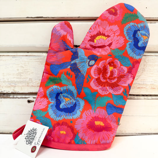 A single oven mitt with a vibrant floral and bird pattern in pink, blue, red, and green, resting on a white wooden surface with a brand tag attached.