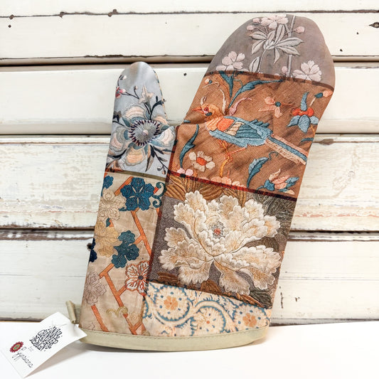 A patchwork quilted oven mitt with floral and bird patterns, resting on a white wooden plank surface with a white tag attached.