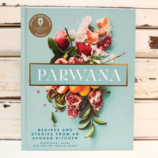 The front cover of the book 'Parwana: Recipes and Stories from an Afghan Kitchen' by Durkhanai Ayubi and Varida Ayubi, featuring a light blue background with a botanical illustration of pomegranates, oranges, and flowers. The book is displayed upright on a rustic white wooden plank surface.