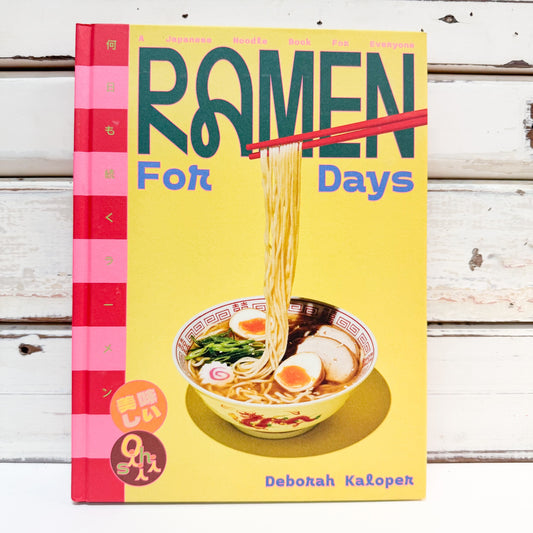 The book 'Ramen For Days' by Deborah Kauperk is shown flat-laid on a white wooden plank surface. The cover is bright yellow with a pink striped spine, featuring a graphic illustration of a bowl of ramen with chopsticks lifting noodles.