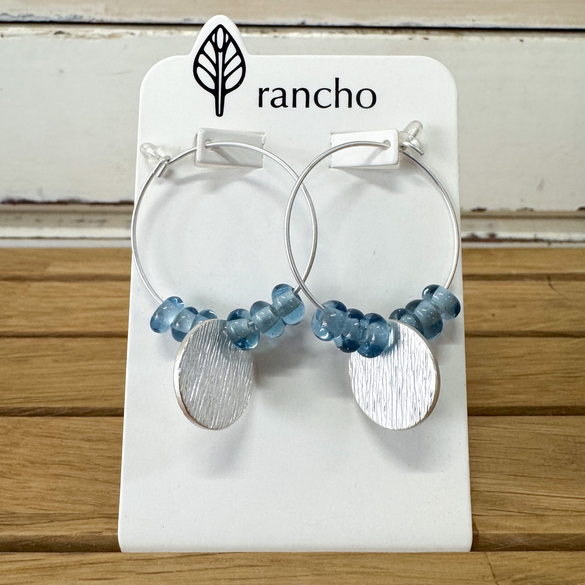Pair of hoop earrings with blue beads and silver discs on a Rancho branded card.Rancho Designs - Blue Beaded Silver Disc Earrings