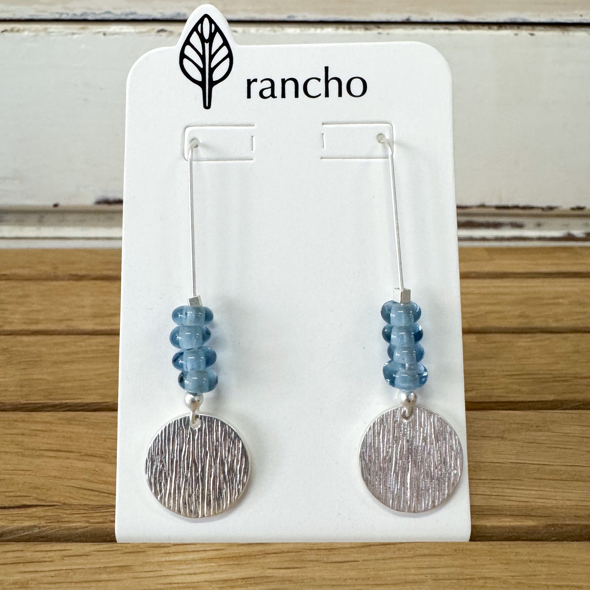 Pair of earrings with blue beads and silver circular ends on a Rancho branded card.
Rancho Designs - Blue Beads Silver Earring