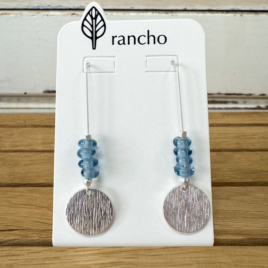Pair of earrings with blue beads and silver circular ends on a Rancho branded card.
Rancho Designs - Blue Beads Silver Earring