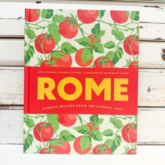 A hardcover cookbook titled 'ROME: Classic Recipes from the Eternal City' featuring a vibrant red and yellow cover design with a green and red tomato pattern, set against a rustic white wooden background.