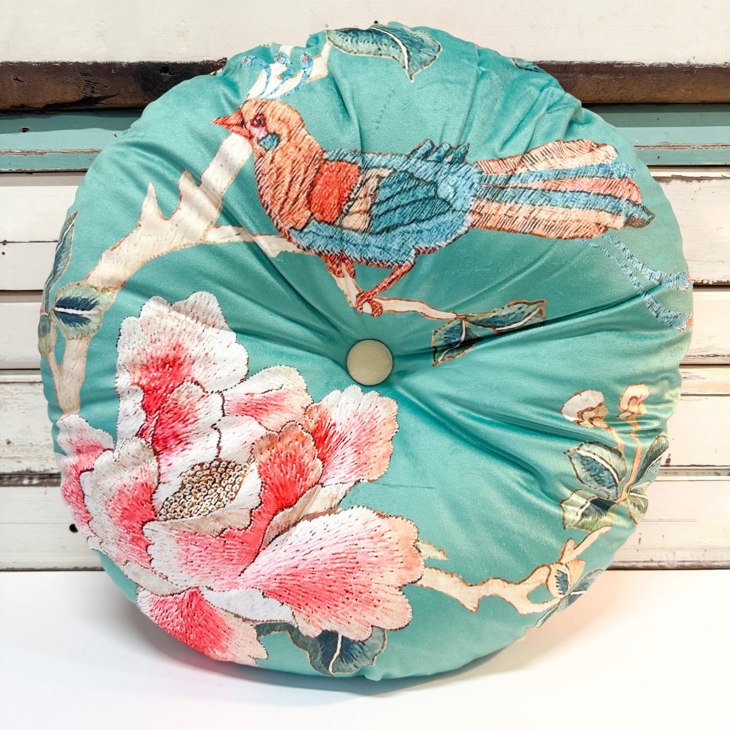 A round tufted cushion with a vibrant turquoise fabric featuring an embroidered bird and large pink and white peony flowers, set against a rustic white wooden background.