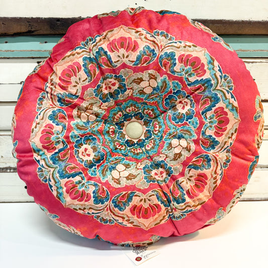A round decorative throw pillow with a vibrant pink border and an intricate, symmetrical mandala-style floral pattern in blue, beige, and brown, displayed against a white wooden background.