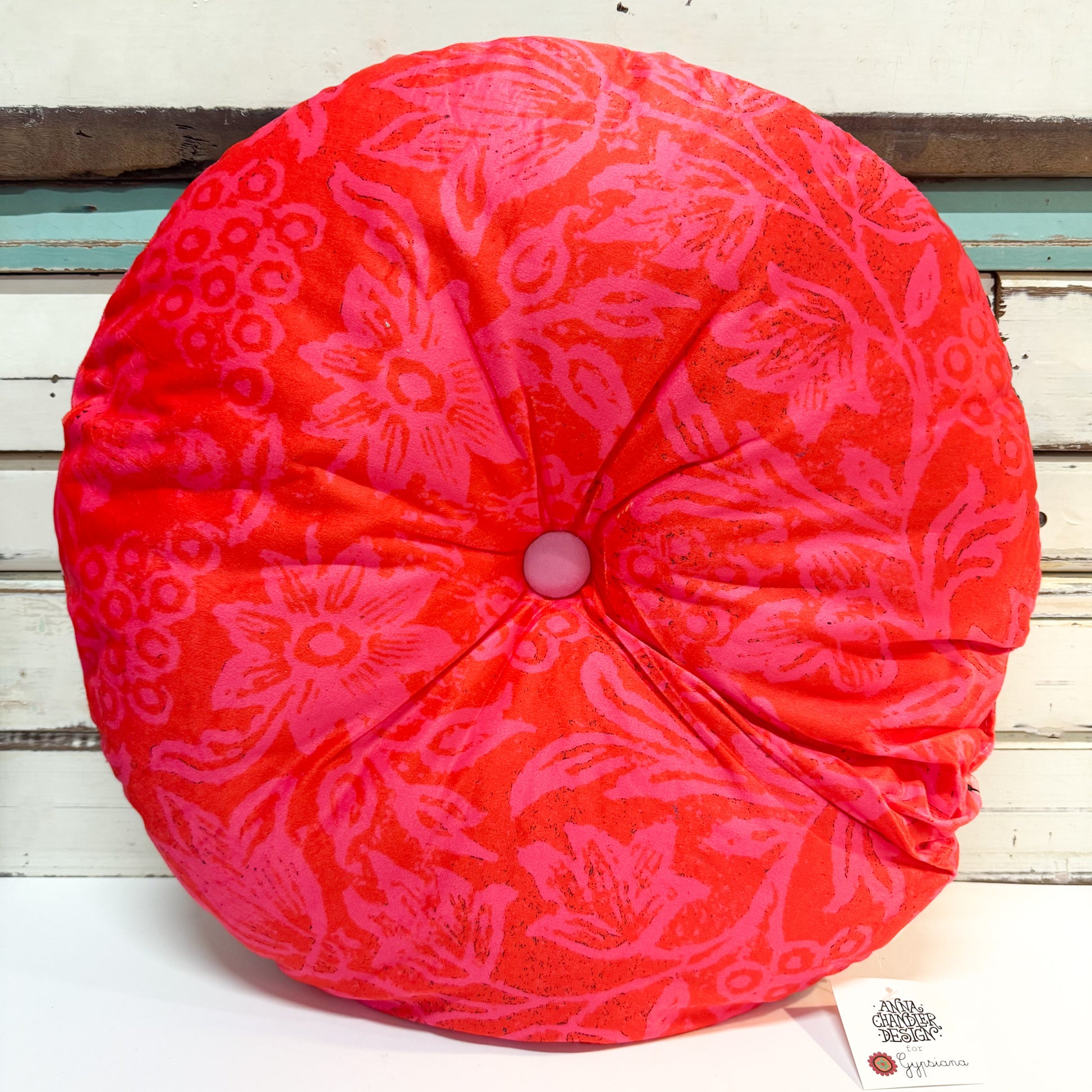 A round, tufted cushion in a vibrant red-orange fabric with a pink floral jacquard pattern, shown against a rustic white-painted wooden background.