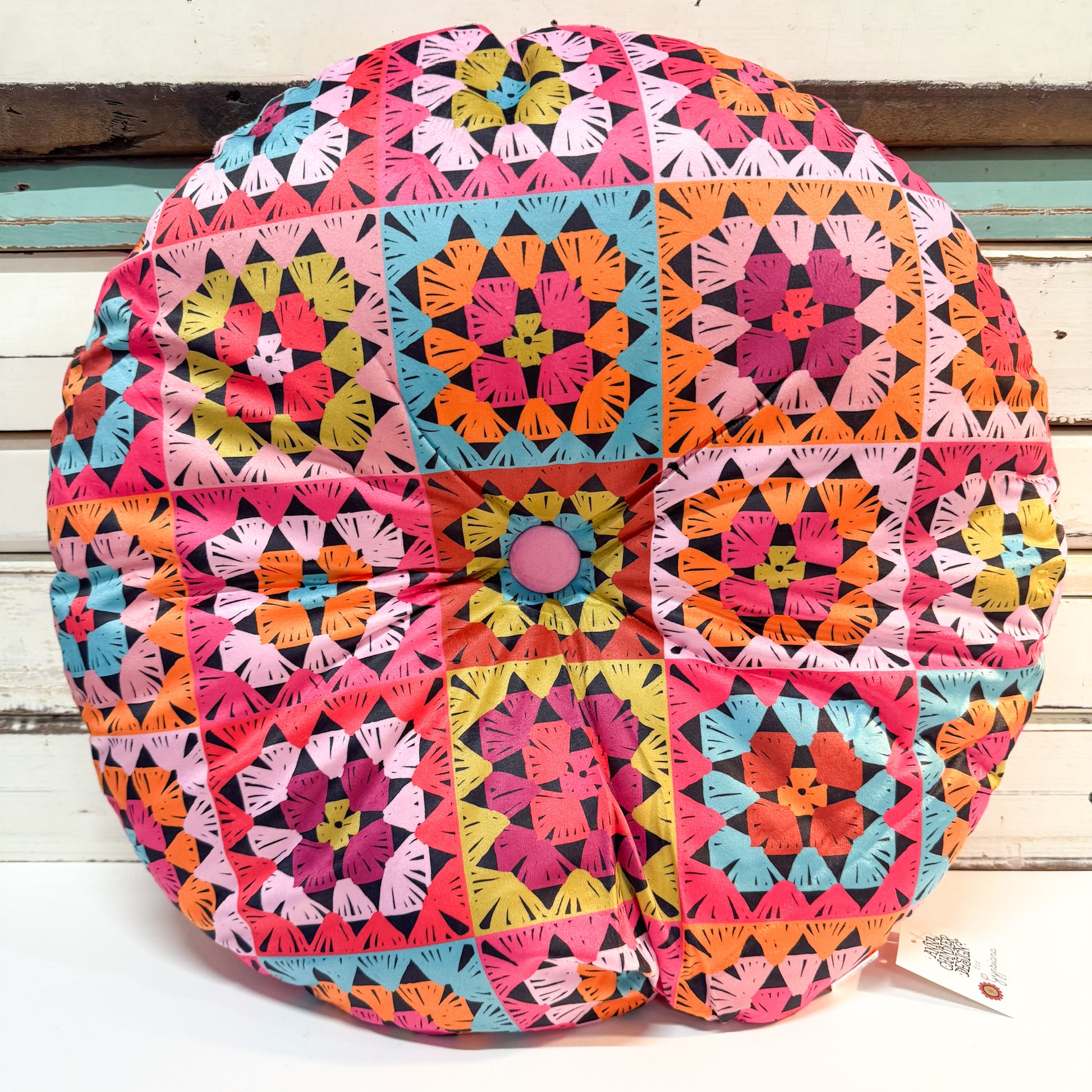 A round decorative throw pillow featuring a vibrant, multicolored patchwork design of geometric quilted squares in pink, orange, blue, yellow, and green, with a central pink button, set against a white wooden background.