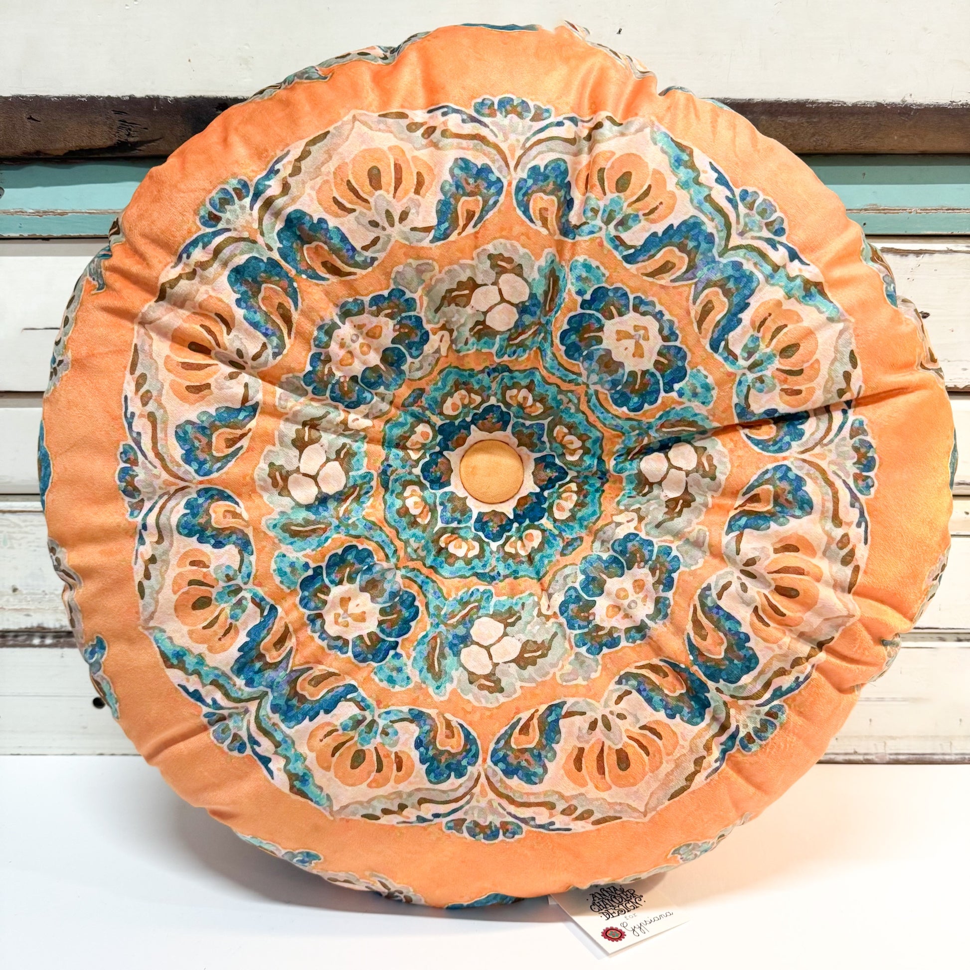 A round decorative throw pillow with a vibrant mandala pattern in orange, teal, blue, and beige, resting on a white wooden surface.