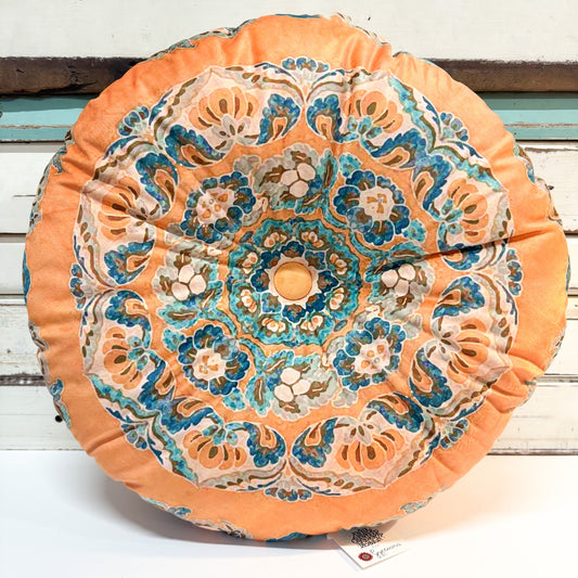 A round decorative throw pillow with a vibrant mandala pattern in orange, teal, blue, and beige, resting on a white wooden surface.