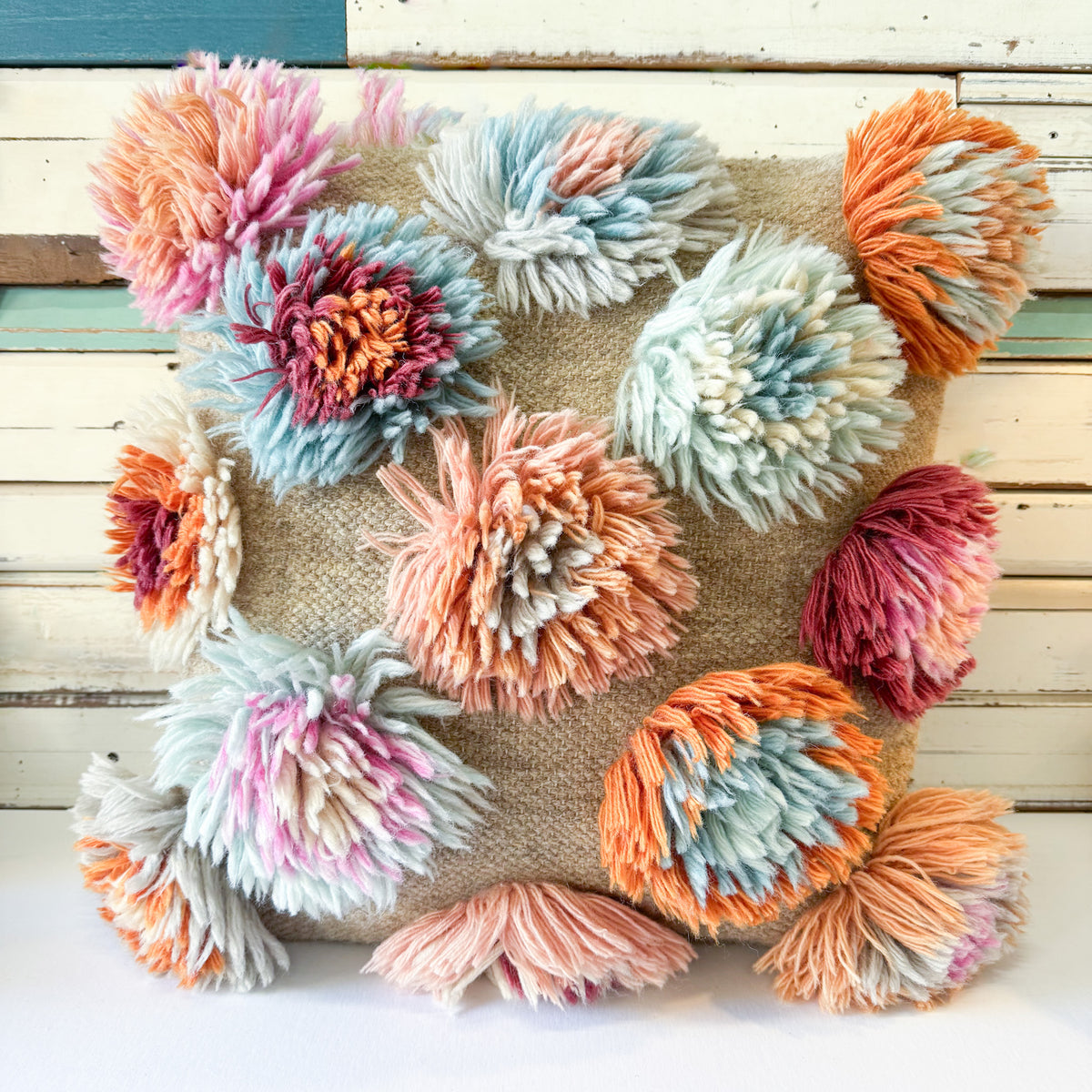 A square decorative throw pillow made from natural burlap fabric, decorated with multiple large, fluffy pom-poms in pastel pink, peach, orange, light blue, and cream, set against a rustic white wooden plank background.