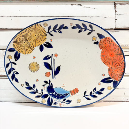 An oval ceramic serving platter with a hand-painted folk art design, featuring blue birds, orange and yellow flowers, and blue leafy vines on a speckled off-white background, set against a rustic white wood plank backdrop.