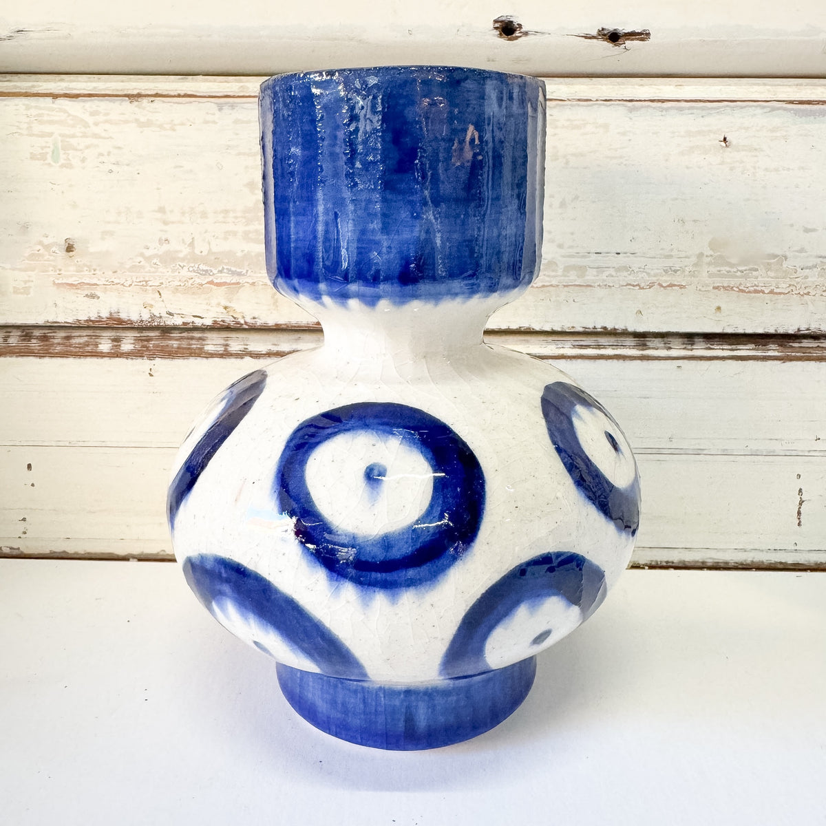 A handmade ceramic vase with a bulbous body and cylindrical neck, decorated with blue and white evil eye patterns. The Brightside Store | Blue Romane Vase - Short