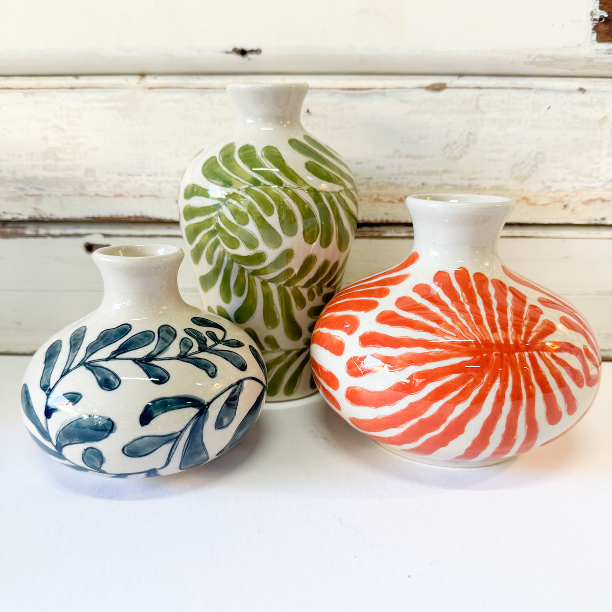 Three small ceramic vases with white backgrounds and colorful painted patterns, displayed on a white surface against a rustic white wooden backdrop.