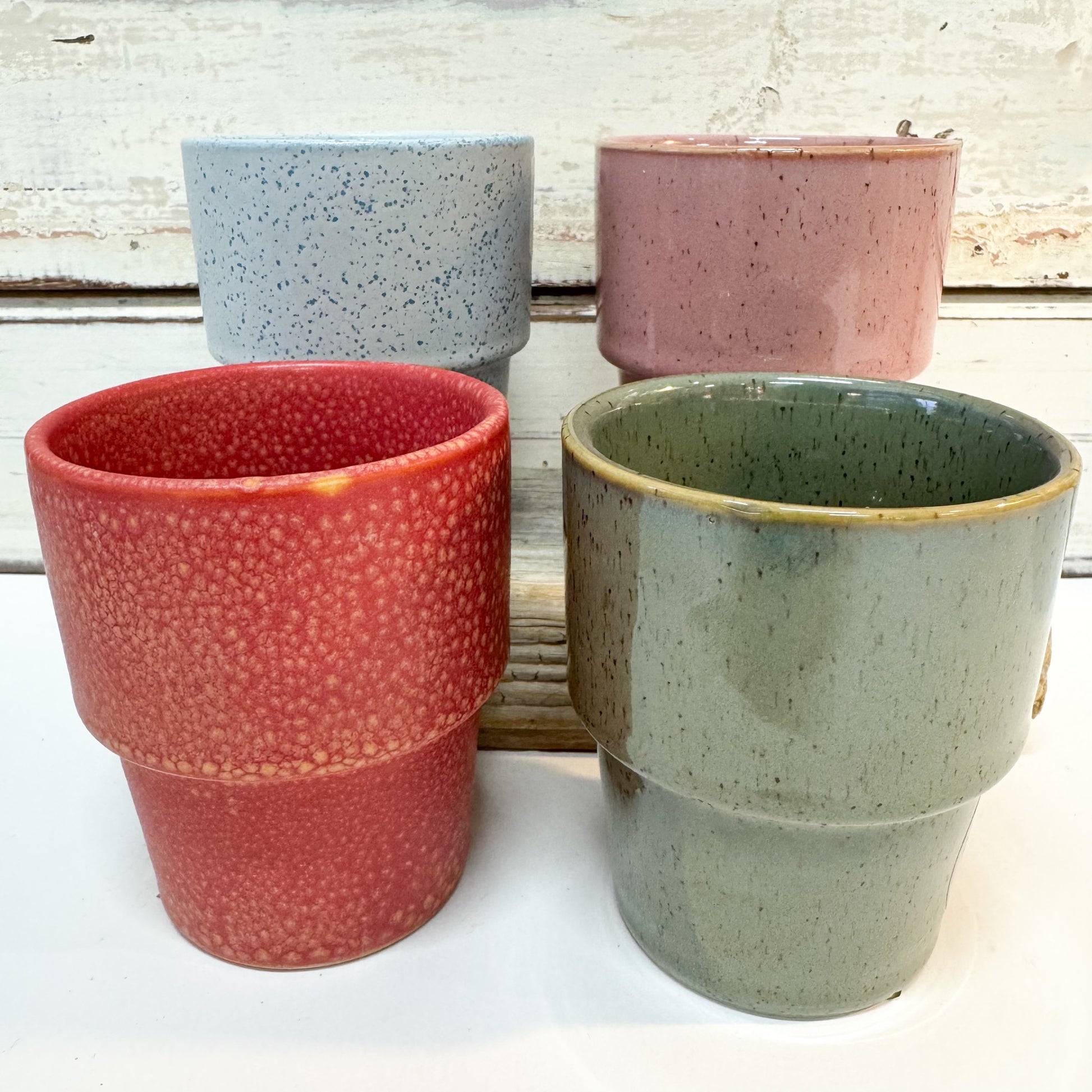 Set of four ceramic cups in different colours and textures, including a red speckled one, a pink speckled one, a light blue speckled one, and a sage green glazed one, 