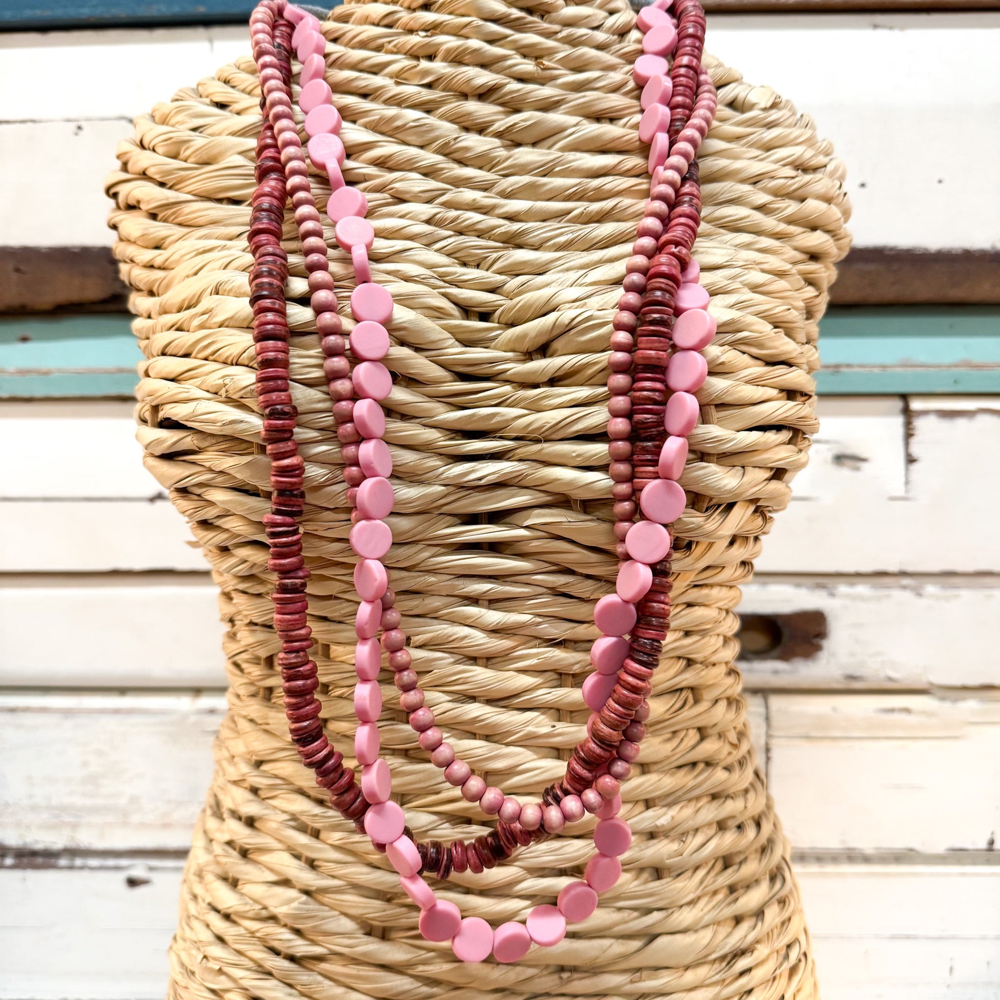 The Brightside Store | Islander Necklace ~ Coral Reef A three-strand beaded necklace draped over a woven straw mannequin torso, featuring alternating strands of dark reddish-brown disc beads and light pink disc beads, set against a rustic white wooden background.