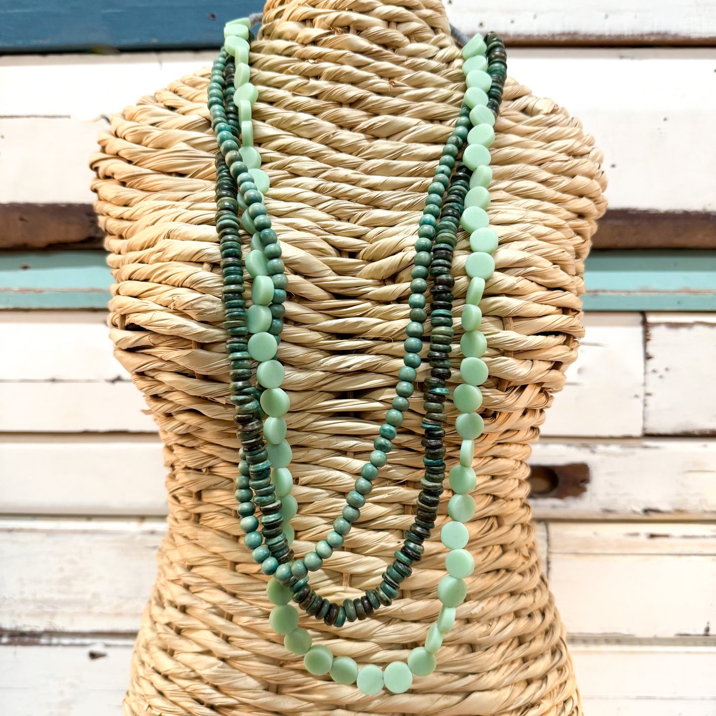 The Brightside Store | Islander Necklace ~ Sea Greens A three-strand beaded necklace set in shades of green and mint, displayed on a woven straw mannequin torso against a rustic white wooden plank background.
