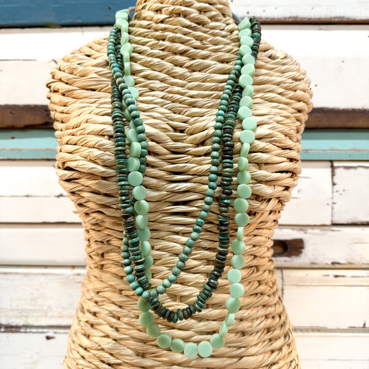The Brightside Store | Islander Necklace ~ Sea Greens A three-strand beaded necklace set in shades of green and mint, displayed on a woven straw mannequin torso against a rustic white wooden plank background.