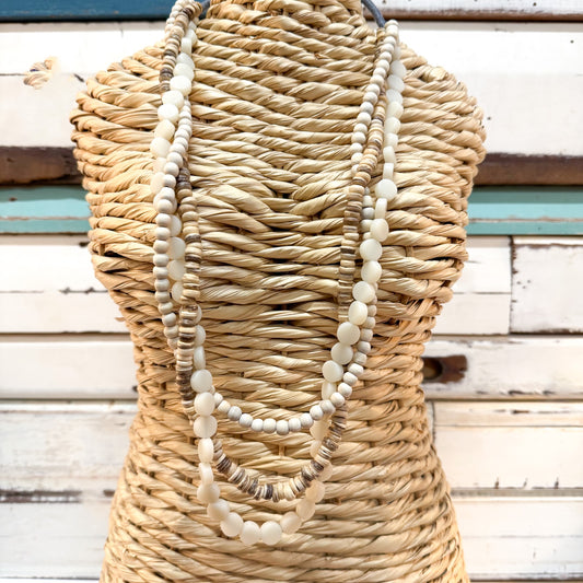 The Brightside Store | Islander Necklace ~ White Sands A three-strand beaded necklace with cream, white, and light brown beads draped over a woven straw mannequin torso, set against a rustic white wooden plank background.