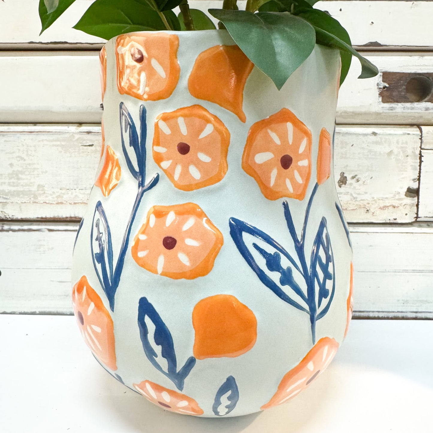 The Brightside Store | Juniper Vase Blue A ceramic planter with a rounded bulbous shape, decorated with hand-painted orange flowers and blue leaves on a white background, holding green plant leaves against a white wooden paneled backdrop.